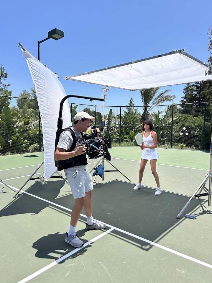 A camera operator films a player on a sunlit court under an overhead diffusion frame.